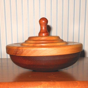 Peut inclure: Un bol en bois artisanal avec un couvercle. Le bol est fait de deux tons de bois, avec une base marron foncé et un couvercle marron clair. Le couvercle a une petite poignée arrondie. Le bol est posé sur une surface en bois.