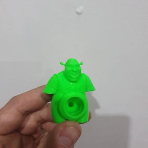 May include: A vibrant green 3D-printed Shrek figurine. The figure features a smiling face, small ears, and a circular opening in the torso. The figurine is held against a plain white background. The figure is approximately 7 cm tall.