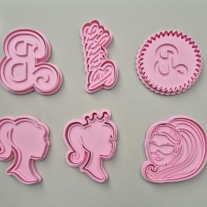 May include: A selection of pink plastic cookie cutters in different shapes. The shapes include the ampersand symbol, the word "Barbie" in cursive, a round shape with a scalloped edge and an ampersand, and three profile silhouettes of a woman.