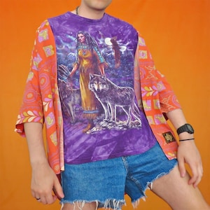 Puede incluir: Una camiseta morada teñida con una mujer nativa americana, lobos y un águila. La camiseta se usa con un kimono naranja estampado y pantalones cortos de mezclilla. Se ve un reloj negro en la muñeca.