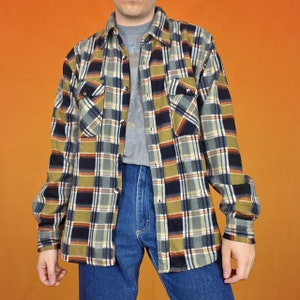 Peut inclure: Une chemise en flanelle à manches longues avec un motif à carreaux dans les tons de vert, noir, orange et blanc. La chemise a un devant boutonné et deux poches poitrine.