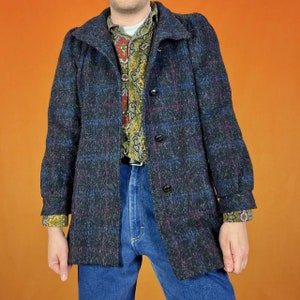 Puede incluir: Un abrigo vintage de color oscuro con un estampado de cuadros en tonos azules, morados y negros. El abrigo tiene una parte delantera abotonada y un cuello. Debajo se lleva una camisa estampada, junto con unos vaqueros azules.