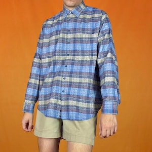 Può includere: Una camicia a maniche lunghe con un motivo a quadri blu, grigio e rosso. La camicia è indossata con pantaloncini color cachi. La camicia ha un colletto classico e polsini con bottoni. Lo sfondo è di colore arancione pieno.