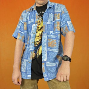 Peut inclure: Une chemise à manches courtes boutonnée à motifs bleus et bruns avec un t-shirt noir en dessous. La chemise a un imprimé graphique d'une personne musclée.