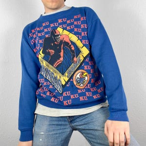 Op de afbeelding: Blauw sweatshirt met een afbeelding van een American footballspeler en de tekst "The University of Kansas Jayhawks". Het sweatshirt is bedekt met de letters "KU" in rood.