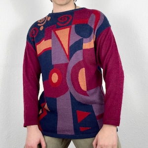 May include: A long-sleeved jumper with a geometric pattern in shades of red, purple, blue, and orange. The jumper has a crew neck and a relaxed fit.