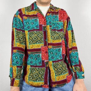 Può includere: Una camicia a maniche lunghe con bottoni e un design a patchwork. La camicia presenta una varietà di colori e motivi, tra cui rosso, giallo, verde, blu e marrone. La camicia ha un colletto e una chiusura con bottoni sul davanti.