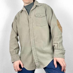 Puede incluir: Una camisa de manga larga de color verde claro con un parche en el codo derecho. La camisa tiene un bolsillo en el pecho y un cuello abotonado.