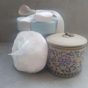 May include: A round, beige ceramic jar with a blue and white floral pattern, a lid with a small metal ring handle, a fluffy white powder puff with a white ribbon, and a light blue gift box with a white satin ribbon tied in a bow on top.