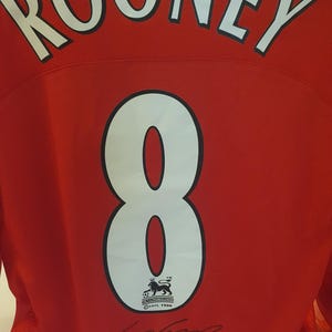 May include: A red football shirt with the name "ROONEY" printed on the back in white. The number "8" is printed in white on the back of the shirt. The shirt has a black and white crest with a lion and the text "The Football Association Premier League" and "FAPL 1996". The shirt is signed in black ink by "Wayne Rooney".