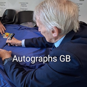 May include: A man in a dark blue suit is signing a blue jersey with a black marker. The jersey has a crest on the left side and the text "Autographs GB" is printed on the bottom right corner.