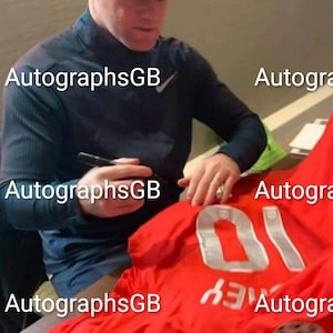 Puede incluir: Una camiseta de fútbol roja con el número 10 en la espalda. La camiseta está siendo firmada por una persona que lleva una camisa gris y una gorra de béisbol negra.