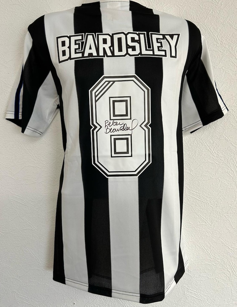 Op de afbeelding: Zwart-wit gestreept voetbal shirt met de naam "BEARDSLEY" en het nummer "8" op de rug. Het shirt is gesigneerd door "Peter Beardsley".