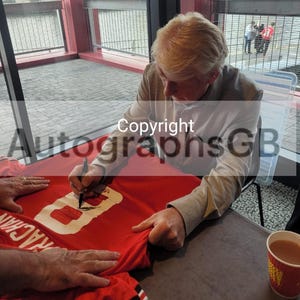 Könnte beinhalten: Eine Person unterschreibt ein rotes Fußballtrikot mit der Nummer 8 in Weiß. Das Trikot hat schwarze Streifen an den Ärmeln. Eine Tasse Kaffee steht auf dem Tisch. Die Wörter "Copyright" und "AutographsGB" sind sichtbar.