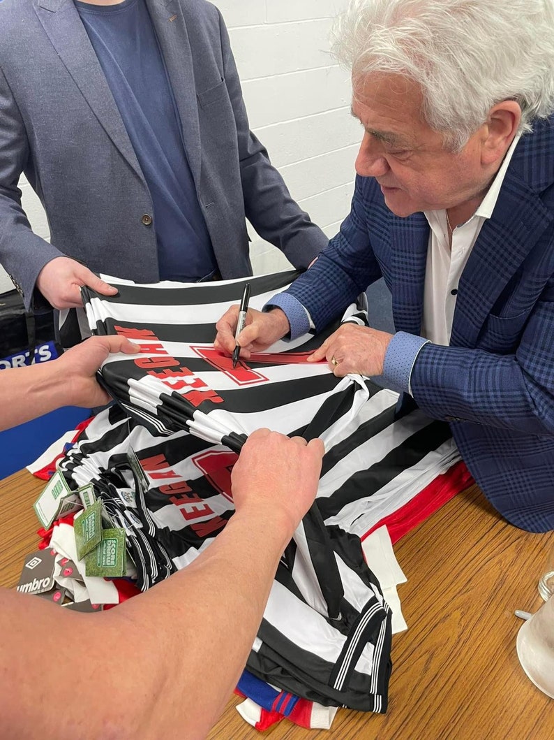 Może przedstawiać: Osoba podpisująca czarno-białą, pasiastą koszulkę piłkarską z czerwonym napisem "Wigan Athletic".