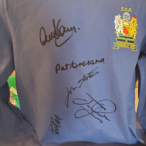 May include: A light blue long-sleeved shirt with multiple signatures in black ink. The shirt features a crest with the text "E.C.F." and "Wembley 1969". The signatures are likely from a sports team.