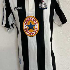 Op de afbeelding: Een zwart-wit gestreept voetbaltenue met het Adidas-logo op de linkerborst en een Newcastle Brown Ale-logo op de voorkant. Het tenue heeft een knoopsluiting.