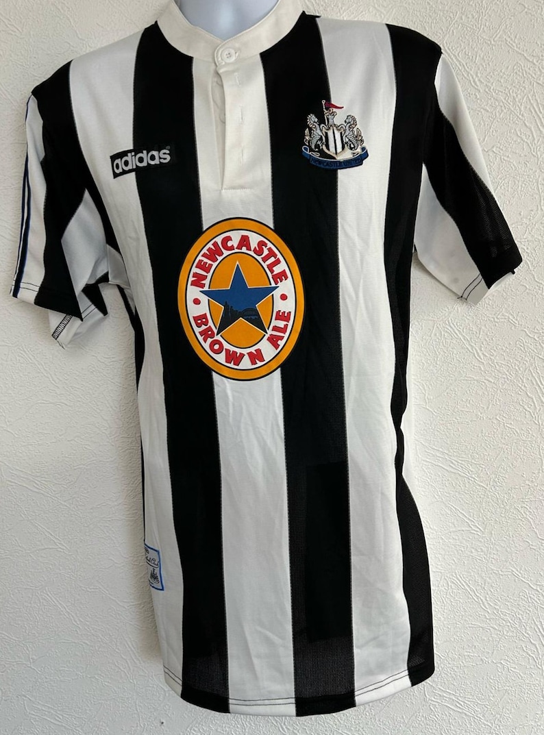 Op de afbeelding: Een zwart-wit gestreept voetbaltenue met het Adidas-logo op de linkerborst en een Newcastle Brown Ale-logo op de voorkant. Het tenue heeft een knoopsluiting.