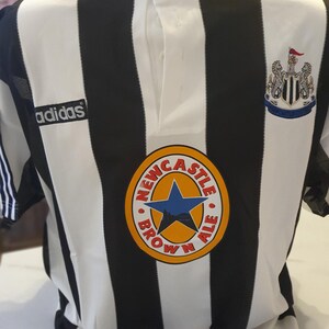 May include: Black and white striped soccer jersey with the Adidas logo on the left chest and the Newcastle Brown Ale logo on the right chest. The jersey has a white collar with a button.