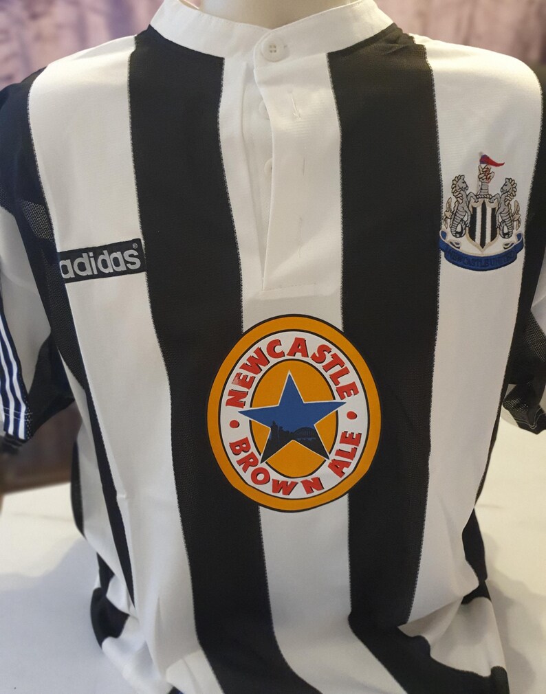 May include: Black and white striped soccer jersey with the Adidas logo on the left chest and the Newcastle Brown Ale logo on the right chest. The jersey has a white collar with a button.