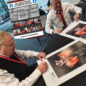 Op de afbeelding: Twee mannen signeren getekende foto's van een voetbalteam dat een trofee vasthoudt. De foto's zijn zwart-wit en hebben een vintage uitstraling. De tekst op het bord luidt "Haal je sportmemorabilia ingelijst met handtekeningen te koop. Gesigneerde sportmemorabilia beschikbaar tegen betaalbare prijzen. Neem gerust een kijkje in mijn Facebook-winkel. Telefoon: 8716882548."