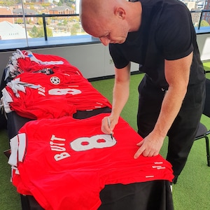 May include: A person signing a red football shirt with the number 8 and the name "Butt" printed on the back.