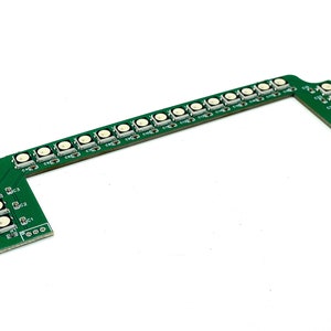 Peut inclure: Une carte de circuit imprimé verte avec plusieurs lumières LED blanches et de petits boutons blancs. La carte a une conception en forme de L avec une série de lumières le long du bras principal et des lumières supplémentaires sur les sections inclinées. Le fond est blanc.