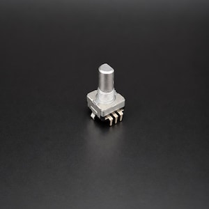 Op de afbeelding: Een zilverkleurige elektronische draaipotentiometer met een cilindrische as met een platte kant, een metalen schroefbus en drie naar beneden gerichte metalen pinnen die uit een vierkante basis steken voor montage op een printplaat.