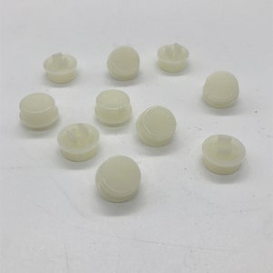 Può includere: Dieci bottoni di plastica bianca con un piccolo centro rialzato. I bottoni sono disposti in cerchio su una superficie bianca.