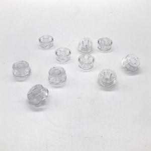 Könnte beinhalten: Eine Gruppe von 11 transparenten Kunststoff-Druckknöpfen mit einem kreuzförmigen Design in der Mitte. Die Knöpfe sind in einem zufälligen Muster auf einer weißen Oberfläche angeordnet.