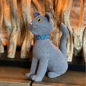 Op de afbeelding: Een grijs gehaakt katten speelgoed met een blauwe kraag. De kat zit met zijn staart om zijn lichaam gekruld. De kat staat voor een houten achtergrond.