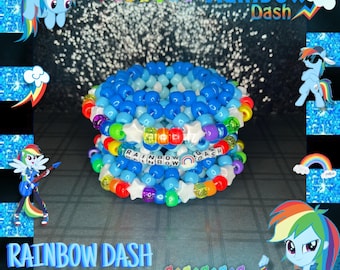 Può includere: Un bracciale a fascia multistrato realizzato con perline di plastica blu, azzurre e bianche. Il design presenta perline bianche a forma di stella e perline con lettere che compongono RAINBOW DASH, con una piccola perlina a tema arcobaleno.
