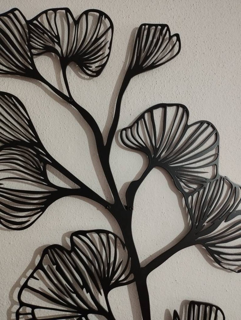 May include: Black metal wall art featuring a stylized ginkgo branch with multiple fan-shaped leaves. The leaves have detailed linear patterns, and the artwork is mounted on a textured white wall. This decorative piece adds a touch of nature-inspired elegance.