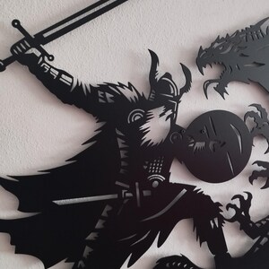 May include: Black metal wall art depicting a warrior with a sword fighting a dragon. The warrior is wearing armor and a helmet with horns. The dragon is attacking the warrior with its claws and teeth.