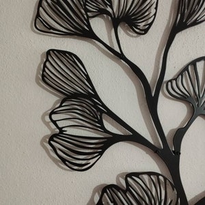 May include: Black metal wall art featuring a stylized ginkgo leaf design. The artwork has a modern, minimalist aesthetic, with intricate lines and a dark finish. The leaves are arranged on slender stems, creating a natural, yet abstract, composition.