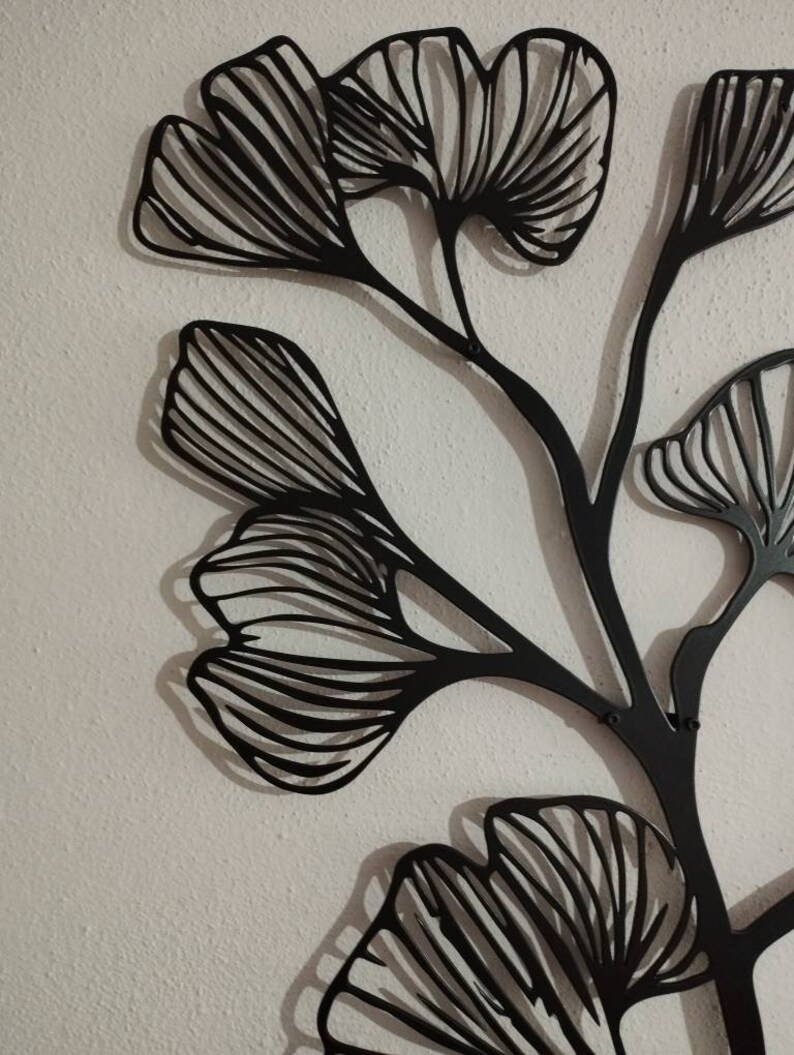 May include: Black metal wall art featuring a stylized ginkgo leaf design. The artwork has a modern, minimalist aesthetic, with intricate lines and a dark finish. The leaves are arranged on slender stems, creating a natural, yet abstract, composition.