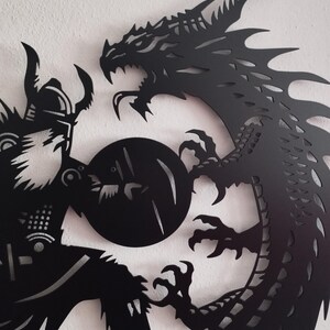 May include: Black metal wall art featuring a silhouette of a dragon and a warrior in combat. The dragon is curled around the warrior, its claws extended. The warrior is holding a shield and a sword.