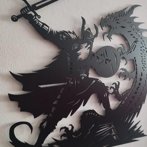May include: Black metal wall art depicting a warrior with a sword and shield fighting a dragon.