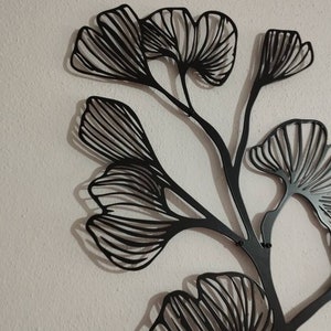 May include: Black metal wall art depicting a stylized ginkgo branch with multiple fan-shaped leaves. The leaves have detailed vein patterns. The artwork is mounted on a textured, off-white wall, creating a contrast between the dark metal and the light background.