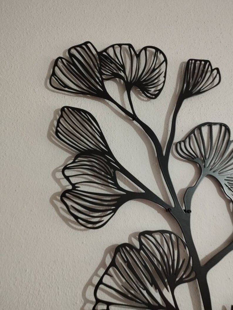 May include: Black metal wall art depicting a stylized ginkgo branch with multiple fan-shaped leaves. The leaves have detailed vein patterns. The artwork is mounted on a textured, off-white wall, creating a contrast between the dark metal and the light background.
