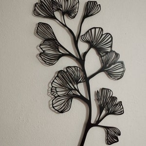 May include: Black metal wall art depicting a stylized ginkgo branch with multiple fan-shaped leaves. The leaves have detailed linear patterns, and the artwork is mounted on a white wall. The design casts a shadow, adding depth to the piece.