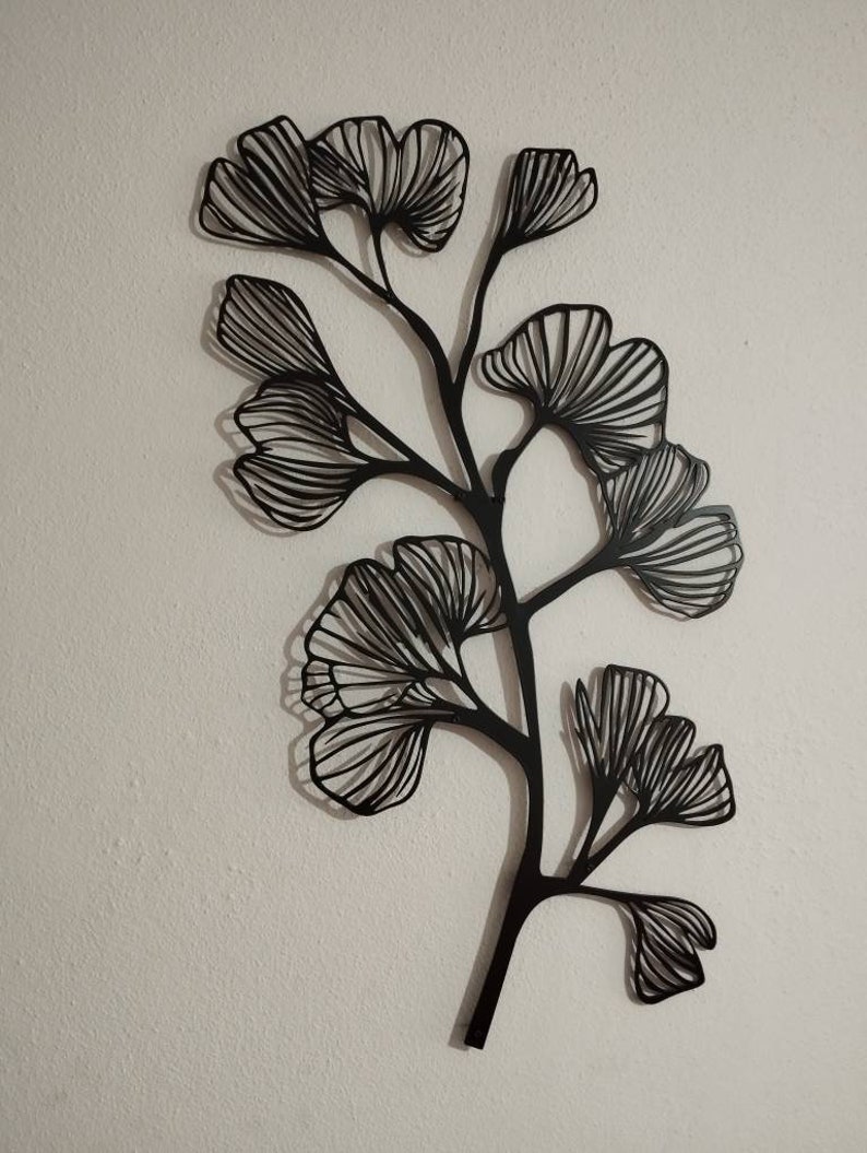 May include: Black metal wall art depicting a stylized ginkgo branch with multiple fan-shaped leaves. The leaves have detailed linear patterns, and the artwork is mounted on a white wall. The design casts a shadow, adding depth to the piece.