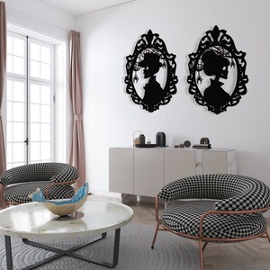 Pode incluir: Duas pe&ccedil;as de decora&ccedil;&atilde;o de parede em silhueta preta em forma de molduras ovais com um perfil de mulher dentro de cada moldura. As molduras s&atilde;o decoradas com desenhos intrincados. A decora&ccedil;&atilde;o de parede est&aacute; pendurada em uma parede branca acima de um arm&aacute;rio branco com duas cadeiras de padr&otilde;es preto e branco na frente.