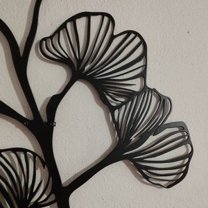 May include: Black metal wall art featuring a stylized ginkgo leaf design. The leaves have a detailed, fan-like structure with visible veins. The art is mounted on a textured, off-white wall, creating a contrast between the dark metal and the light background.