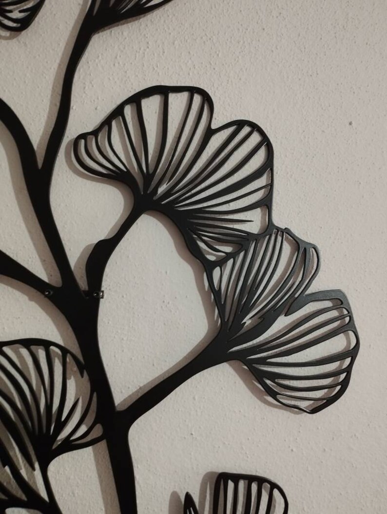 May include: Black metal wall art featuring a stylized ginkgo leaf design. The leaves have a detailed, fan-like structure with visible veins. The art is mounted on a textured, off-white wall, creating a contrast between the dark metal and the light background.