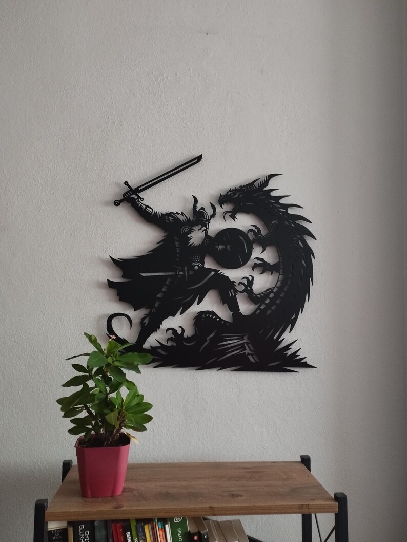 May include: Black metal wall art depicting a Viking warrior battling a dragon. The warrior is holding a sword and shield, while the dragon is attacking with its claws and teeth.