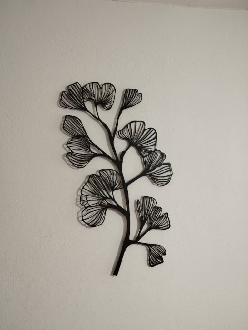 May include: Black metal wall art depicting a stylized ginkgo branch with multiple fan-shaped leaves. The leaves and stems are outlined with thin lines, creating a delicate, airy appearance. The artwork is mounted on a white wall.
