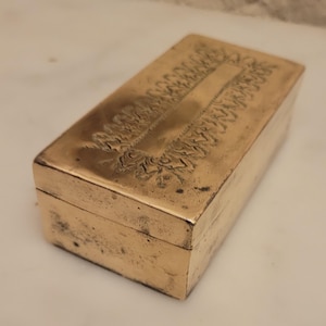 May include: An antique brass box with a hinged lid. The rectangular box has engraved floral patterns on the top and sides. The brass has a warm, aged patina, indicating its vintage. The box is likely for storing small items, such as jewellery.