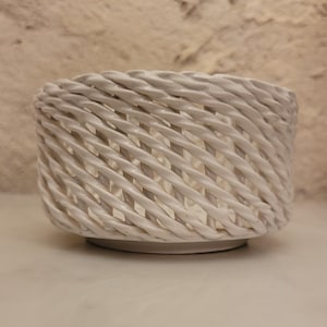 May include: A white ceramic bowl featuring a decorative openwork design that mimics the appearance of braided rope. The structure consists of interconnected, diagonal strands forming a lattice pattern around the cylindrical body of the vessel.