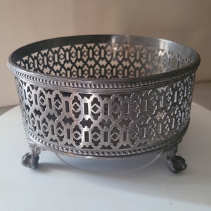 May include: Silver-plated footed bowl with an intricate geometric design. The bowl has a round shape and three legs.